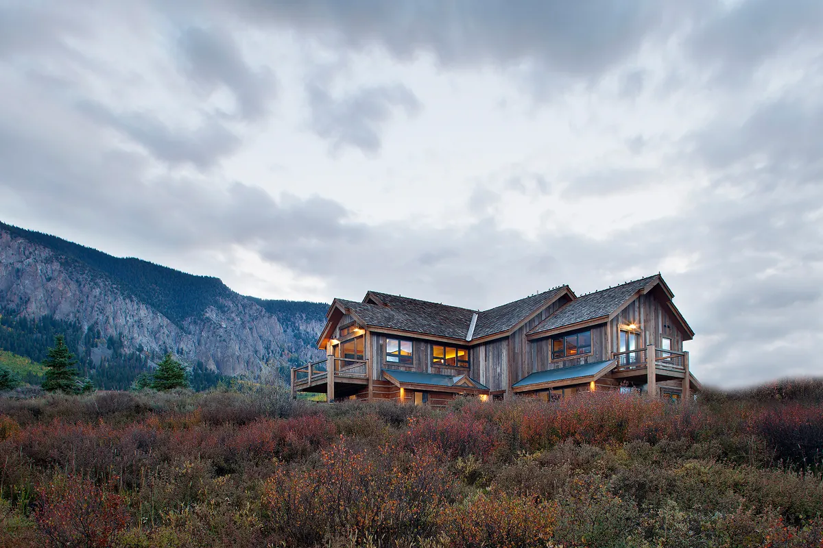 Crested Butte home exterior with reclaimed wood siding, mountain backdrop, and warm interior glow.